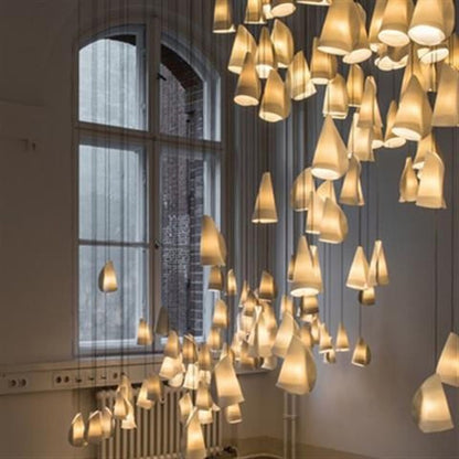Cascading cluster of warm-lit folded bone-white conical pendant shades suspended in front of a tall arched window