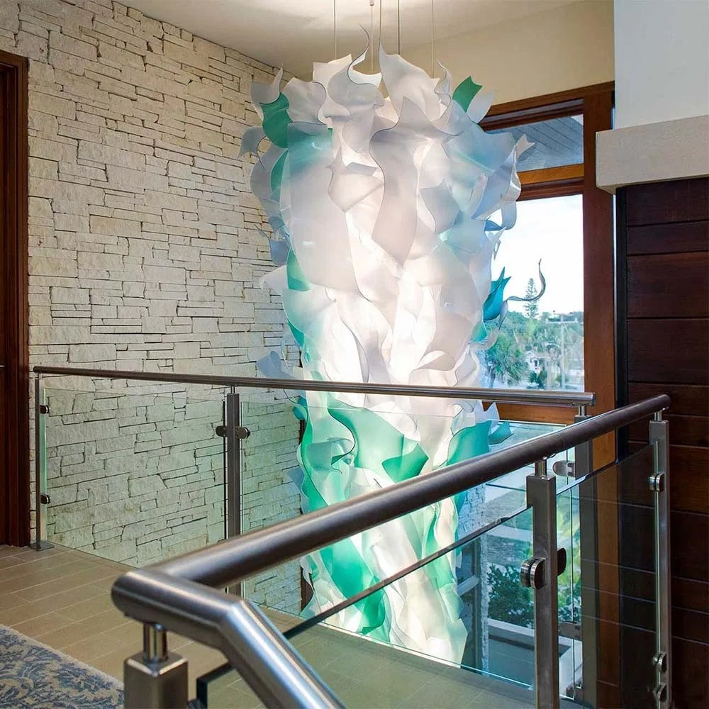 Tall vertical column chandelier of frosted white and seafoam-green bent wave ribbons suspended in a stone-walled stairwell