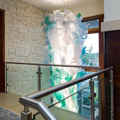 Tall vertical column chandelier of frosted white and seafoam-green bent wave ribbons suspended in a stone-walled stairwell
