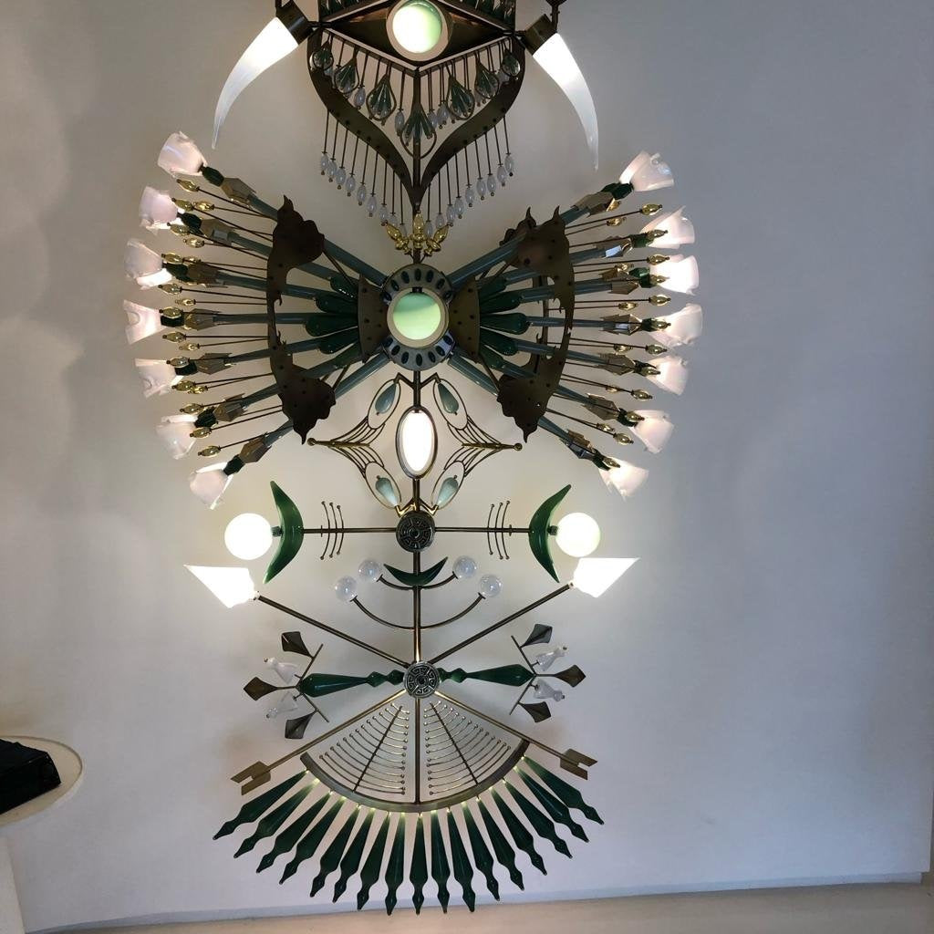 Vertical sculptural totem chandelier in brass, emerald-green glass and illuminated frosted glass shades mounted against a tall wall