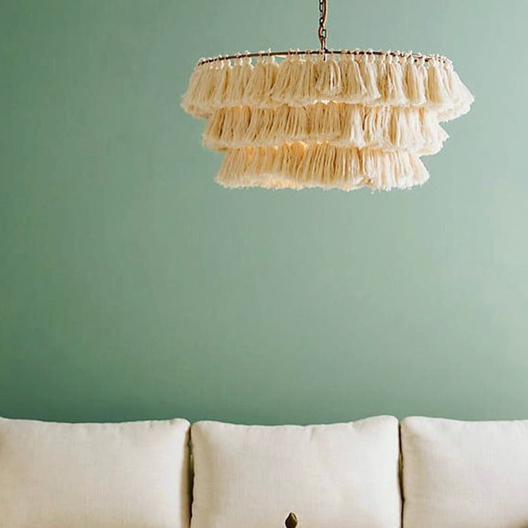 Three-tier cream cotton tassel fringe chandelier suspended on a brass chain against a sage green wall above a cream sofa