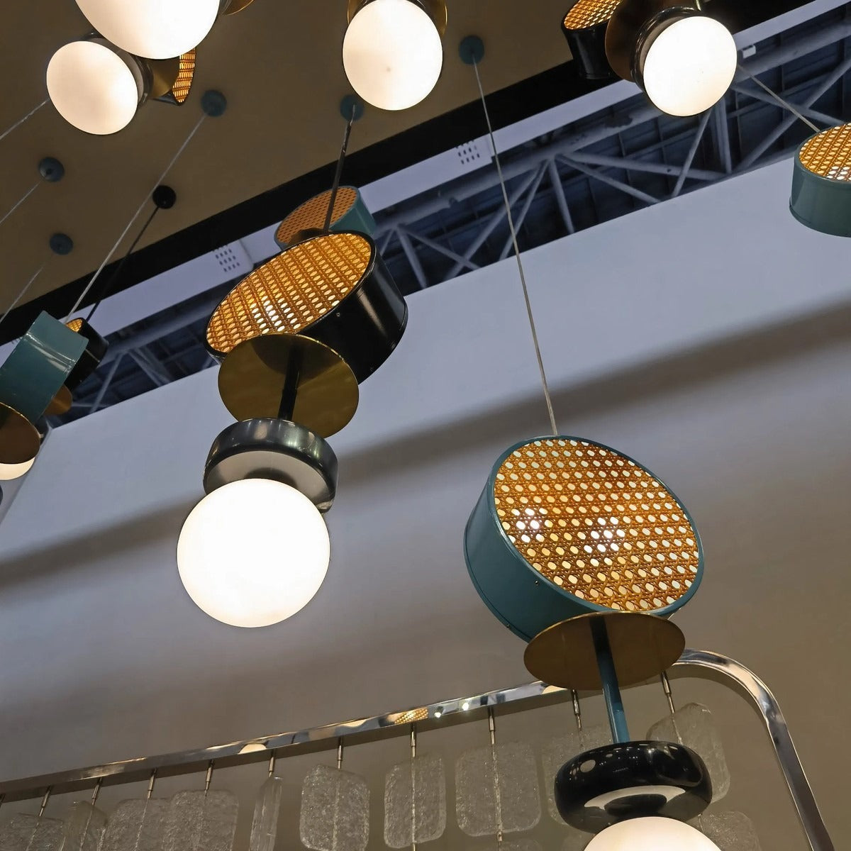 Low-angle close-up of suspended slate-teal and black woven-mesh drum pendants with brass discs and glowing white glass globes