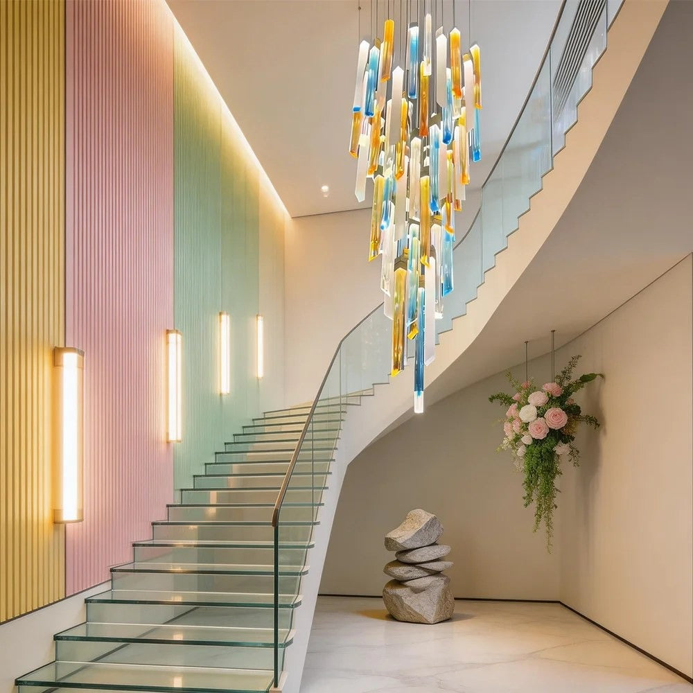 Tapered double-height chandelier of amber, ice-blue, and white vertical art glass bars suspended beside a glass staircase