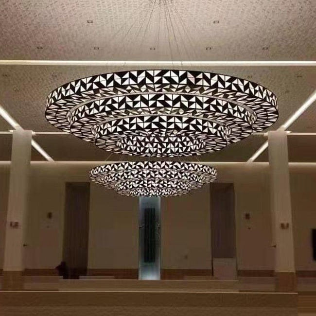 Front view of a four-tier geometric dark bronze lattice ring chandelier with backlit faceted glass panels suspended in a grand hall