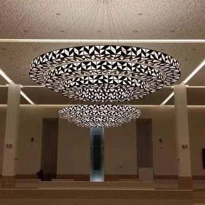 Front view of a four-tier geometric dark bronze lattice ring chandelier with backlit faceted glass panels suspended in a grand hall