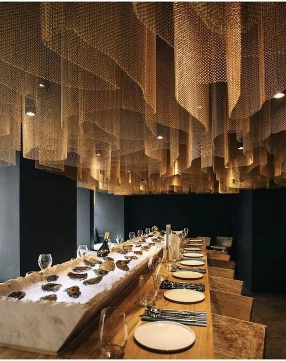 Layered undulating ceiling curtain chandelier made of fine gold-toned metal chains illuminated by warm spotlights over a long dining table