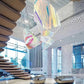 Wide view of the multi-panel iridescent mesh chandelier hanging beside a modern floating wooden staircase in a marble lobby