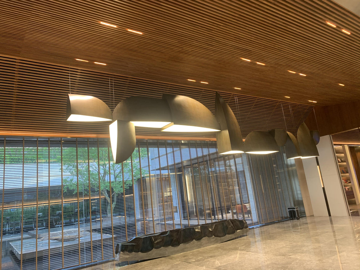 Bronze sail-form pendant chandelier with curved metal panels staggered along a wood-panelled reception ceiling